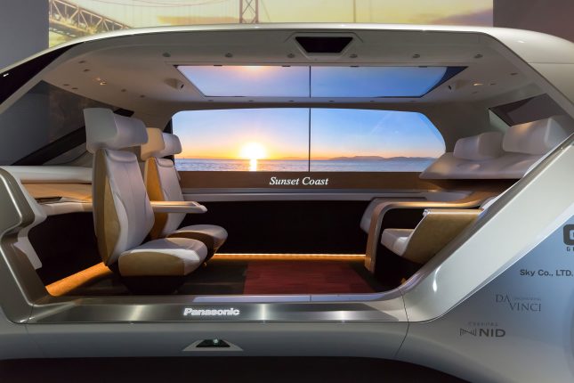 The interior of a panasonic concept vehicle with four light beige seats facing each other and a video screen with a sunset in the background.