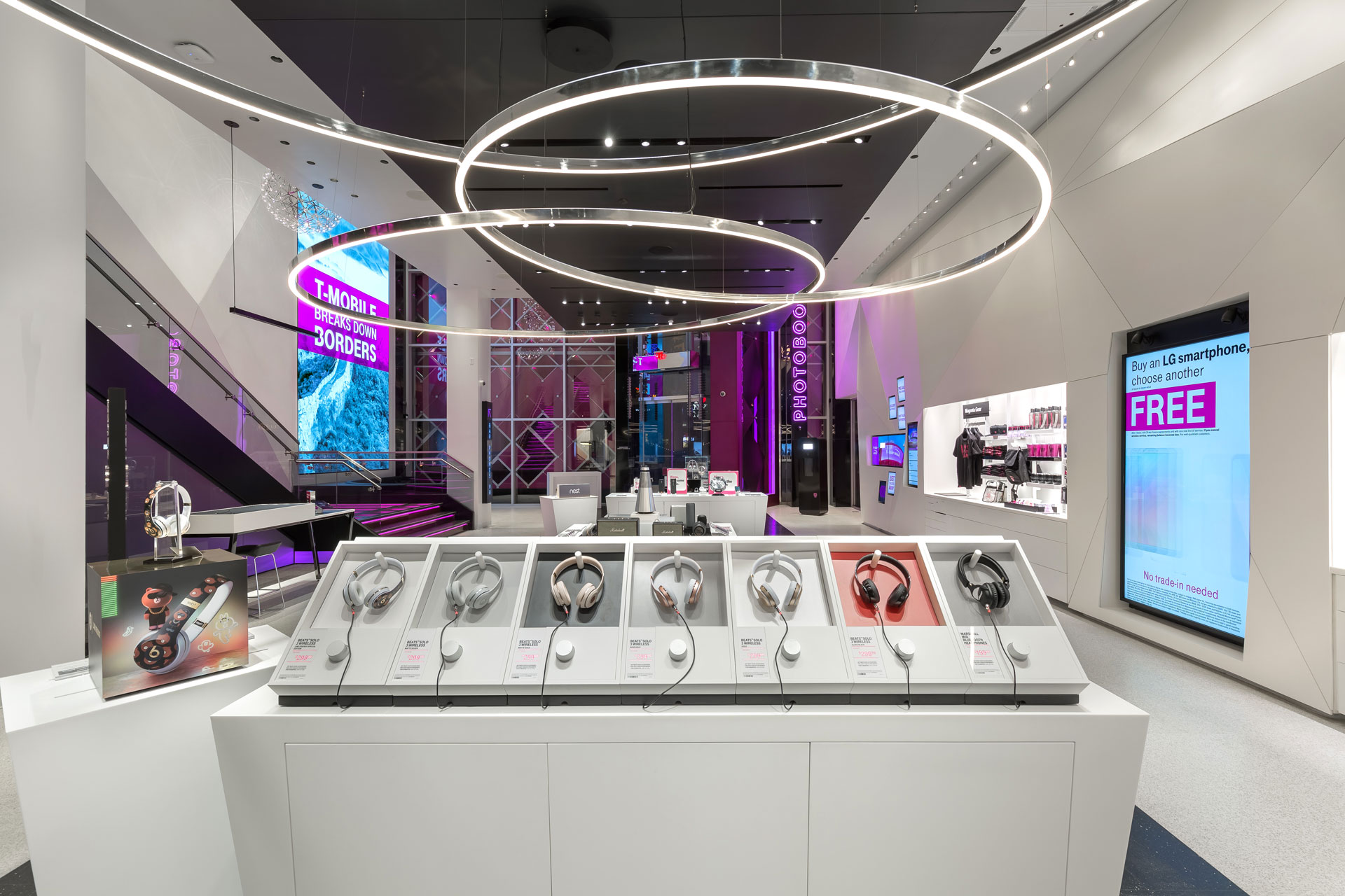 A modern white T-mobile shop interior with a color-coded headphone display int he foreground, thin chrome-sided ring chandeliers above and neon lights in the background.