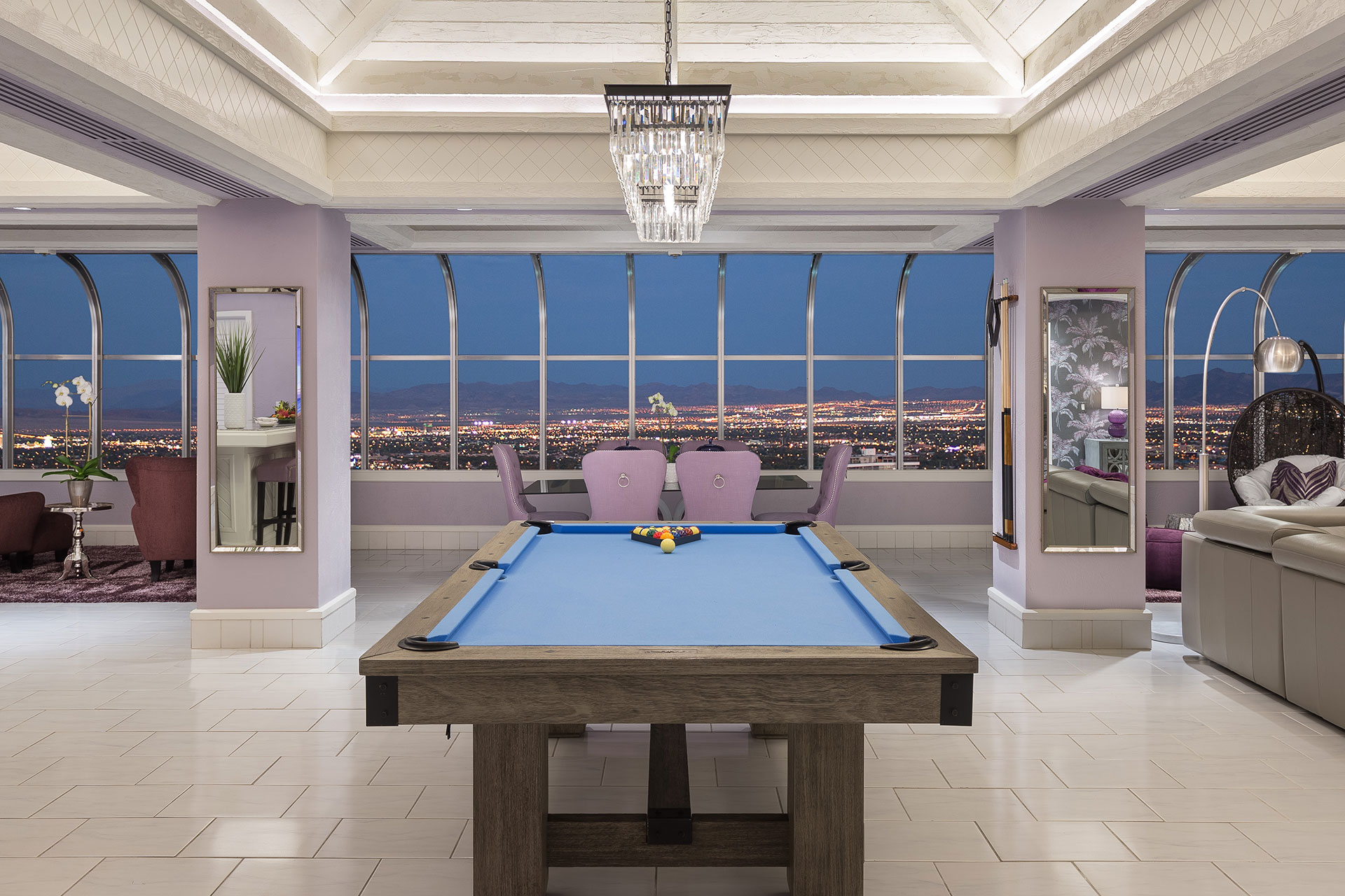 A blue-surfaced pool table in a lavendar-toned hotel room with a dining table and wide city view at dusk in the windows.