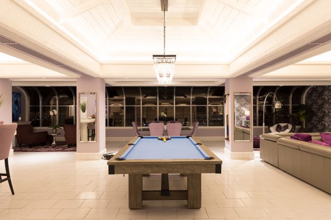 A blue-surfaced pool table in a hotel room withamber color temperature, black windows reflecting glare from the room in the background