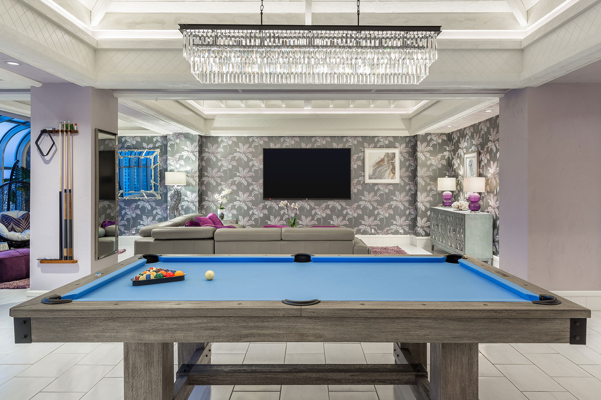 A hotel room with a blue-surfaced pool table in the foreground and a living room with flat-panel TV on silver, patterned wallpaper and a crystal chandelier above.