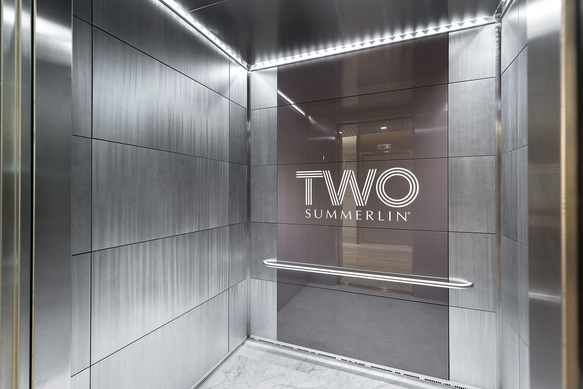 And elevator interior with shiny silver walls and a grey glass center panel with the words "Two Summerlin" written in white and white LED lighting atop.