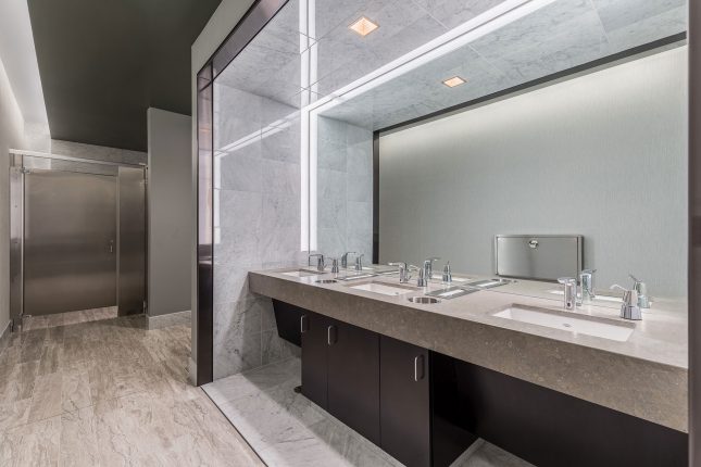 An office building restroom with beige and white tile floor and wall and a beige synthetic countertop with three basins in the foreground and a stainless steel stall door in the background.