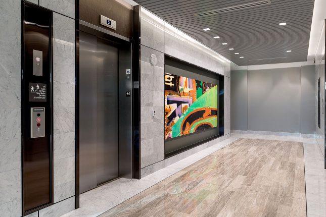 An office building elevator lobby with shiny tile floors and walls and a green, orange, black, and purple piece of abstract art hanging alongside a stainless steel elevator door.