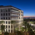 An off-centered perspective of a six-story office building at twilight with beige and grey exterior, three glass balconies, with palm trees and desert lanscaping in the foreground with rich blue skies and the Las Vegas Strip in the background.