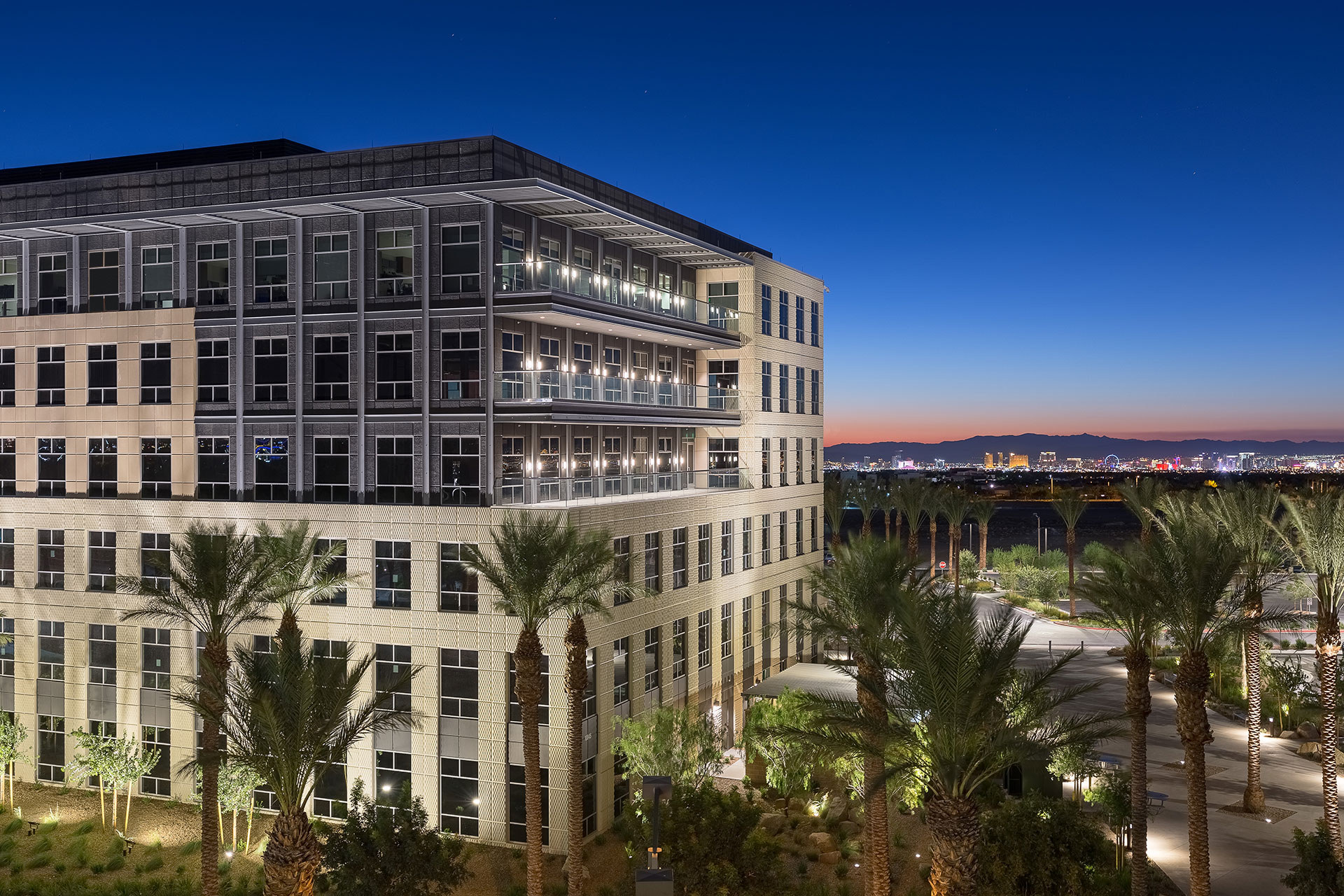 An off-centered perspective of a six-story office building at twilight with beige and grey exterior, three glass balconies, with palm trees and desert lanscaping in the foreground with rich blue skies and the Las Vegas Strip in the background.