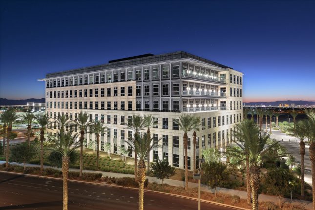 A six-story office building at twilight with beige and grey exterior, three glass balconies, with palm trees and desert lanscaping in the foreground with rich blue skies and the Las Vegas Strip in the background.