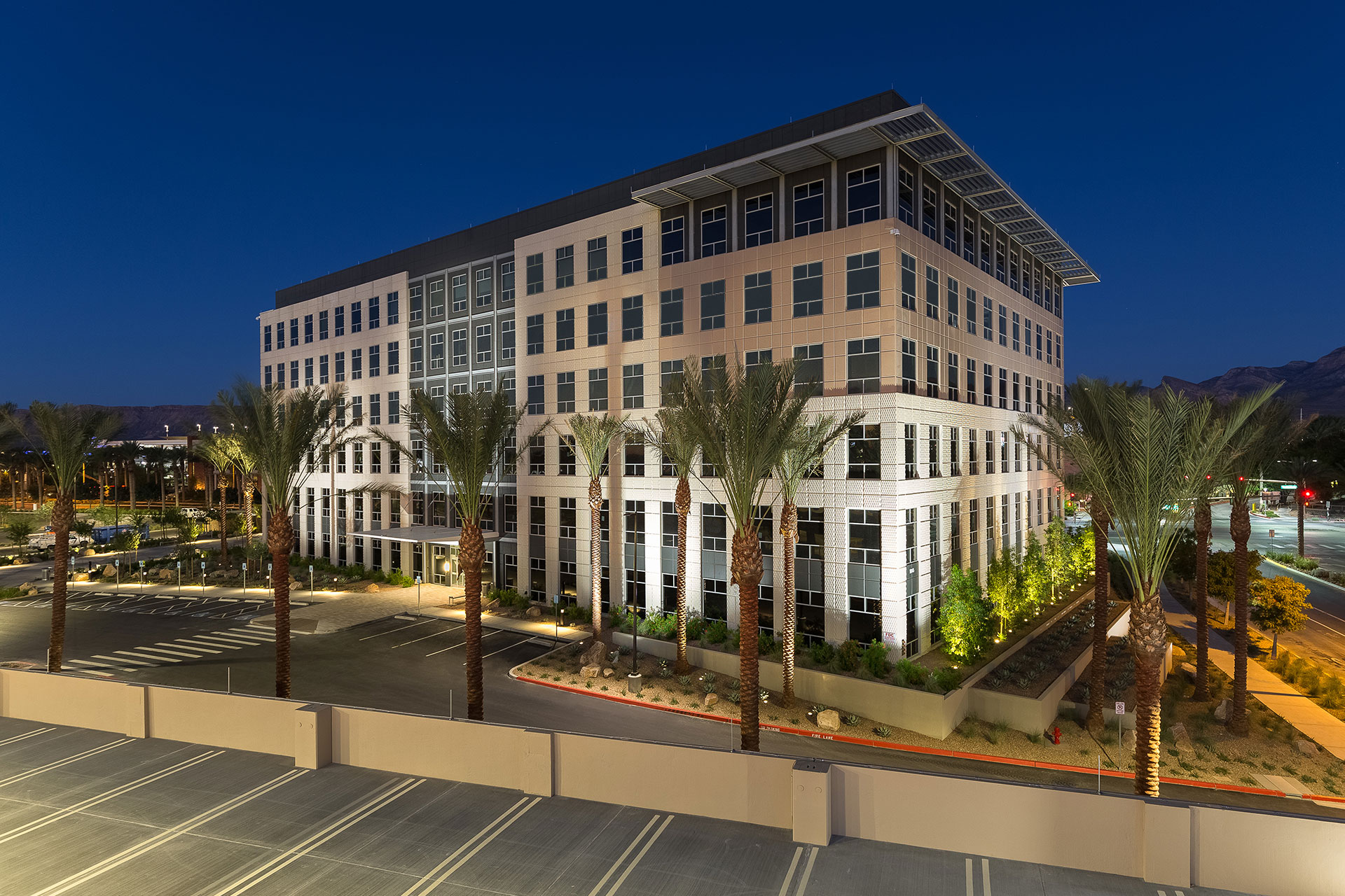 A six-story office building at twilight with beige and grey exterior, with palm trees and desert lanscaping and the top of a parking structure in the foreground with rich blue skies and mountains the background.