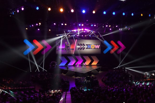 A high-angle view of a packed arena and a stage with spotlights shining up from both sides of the stage and blue, red, and purple lighting and glowing arrows decorating the backdrop with "Next is Now" written on-screen above the stage.