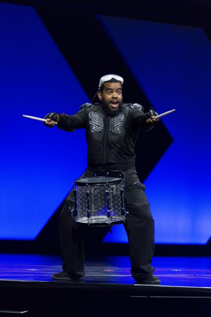 A black man with white goggles on his head dressed in black holding out two drumsticks and wearing a single drum around his waist on a stage with a blue and black background.