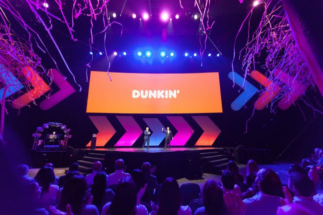 Two men in suits on an arena stage before an audience with blue and purple stage light and confetti flying in from the egdes of the frame and an orange screen with the words DUNKIN' in white in the back.