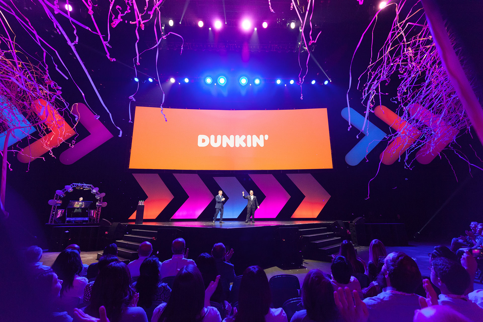 Two men in suits on an arena stage before an audience with blue and purple stage light and confetti flying in from the egdes of the frame and an orange screen with the words DUNKIN' in white in the back.