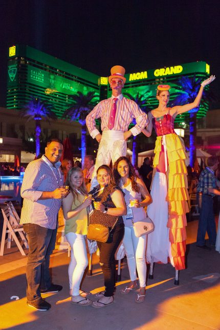 Party-goers posing with entertainers on stils poolside at night with the green MGM Grand hotel in Las Vegas in the backgournd.