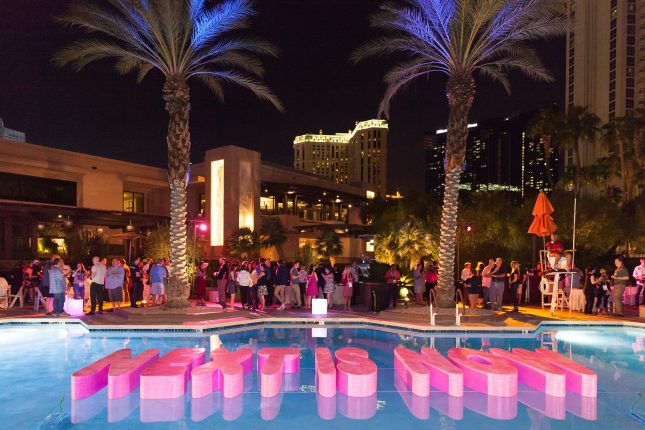 a swimming pool with two large palm trees behind it and pink floating letters spelling NEXT IS NOW floating in it with party-goers around it and hotel buildings in the background.