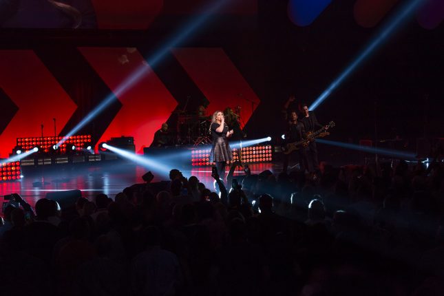 An oblique angle of the stage at a Kelly Clarkson concert with white spotlights shining about and a red and black background.