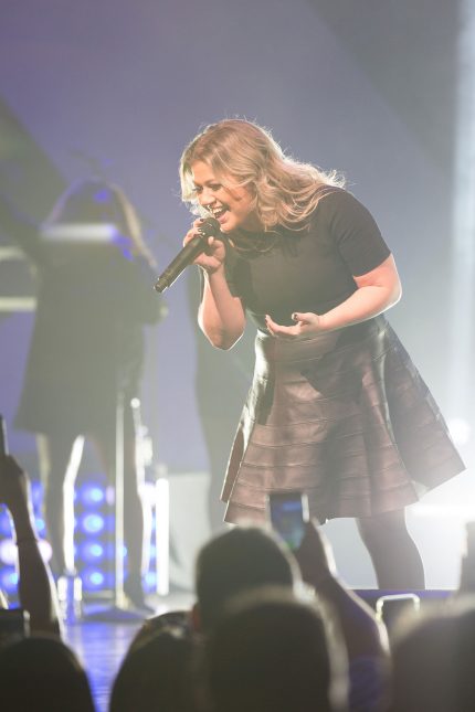 A tight shot of Kelly Clarkson in a black short-sleeved shirt and leather skirt singing into a black handheld microphone on stage in front of a live audience with backup singers and blue and white stage lighting in the background.