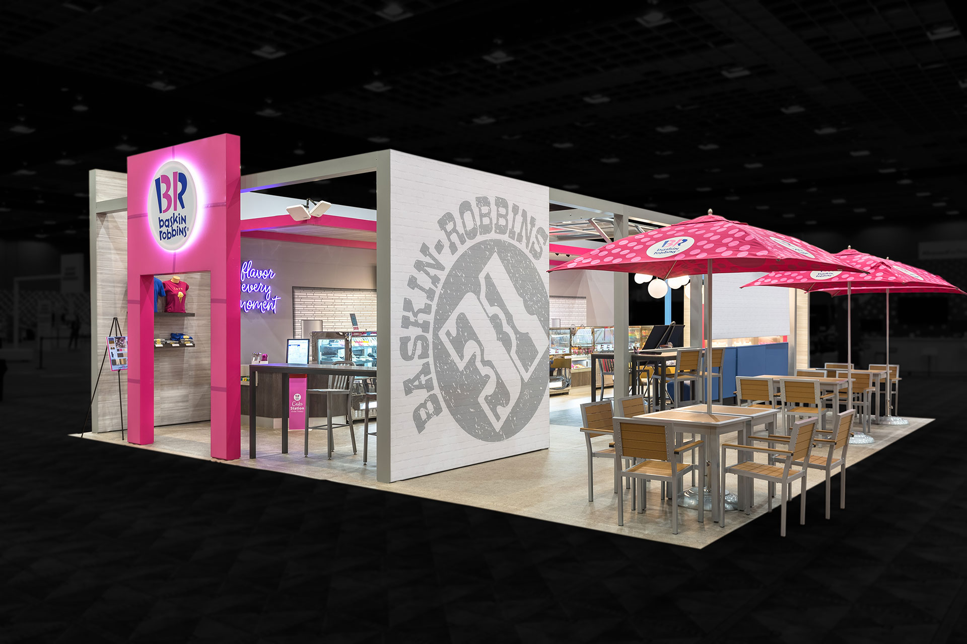 A Baskin Robbins store as a trade show exhibit with a pink arch and logo screen left, a white wall with grey printed logo centered, and tables with pink umbrellas screen right and the interior display cases and counters visible.