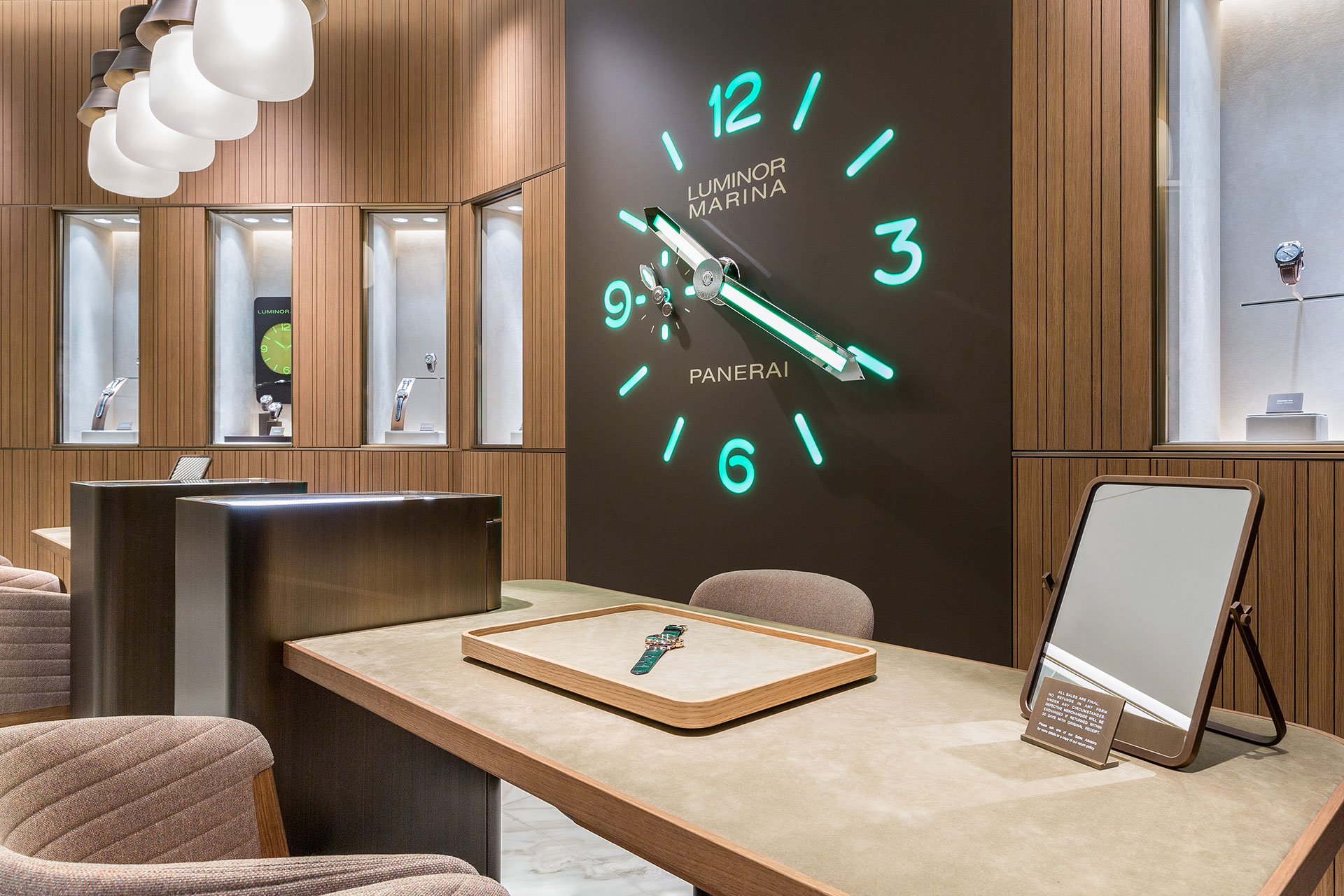 A luxury watch shop with a beige velvet-surface fitting desk in the foreground and a large wall clock in the background with illuminated green hands and numbers beside wood-panel walls with display cases imbedded. 