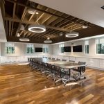 A modern conference room with rich brown hardwood floors, a brown conference table with dark office chairs surrounding it and a long white synthetic countertop above glossy white cabinets surrounding the room and five large flat-panel tvs hung on the walls beside many windows exposing trees outside.