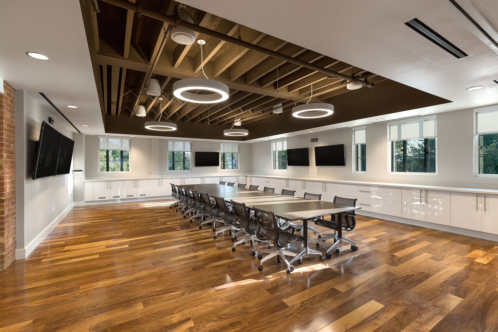 A modern conference room with rich brown hardwood floors, a brown conference table with dark office chairs surrounding it and a long white synthetic countertop above glossy white cabinets surrounding the room and five large flat-panel tvs hung on the walls beside many windows exposing trees outside.