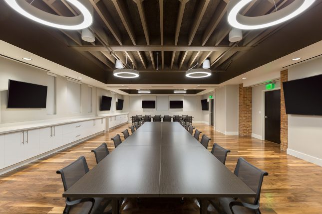 A centered shot of a conference room with rich brown hardwood floors, a vertically-oriented brown conference table in the foreground and a horizontal table in the background with dark office chairs surrounding them and a long white synthetic countertop above glossy white cabinets on the left wall and seven large flat-panel tvs hung throughout and white shades covering multiple windows.