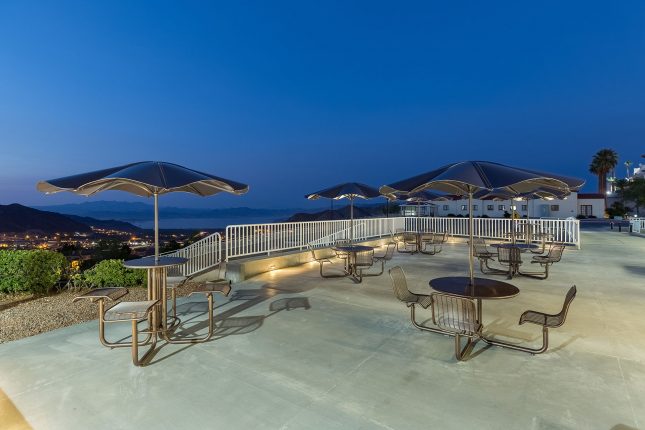 A concrete patio with multiple one-piece metal patio umbrellas with attached table and chairs at dusk with mountains, town lights, and lake in the background under a dark blue sky.