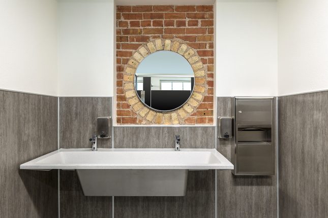 A restroom sink with an exposed red and yellow brick vanity and round mirror, with stainless steel soap and toewl dispensers and a white speckled two-person sink.