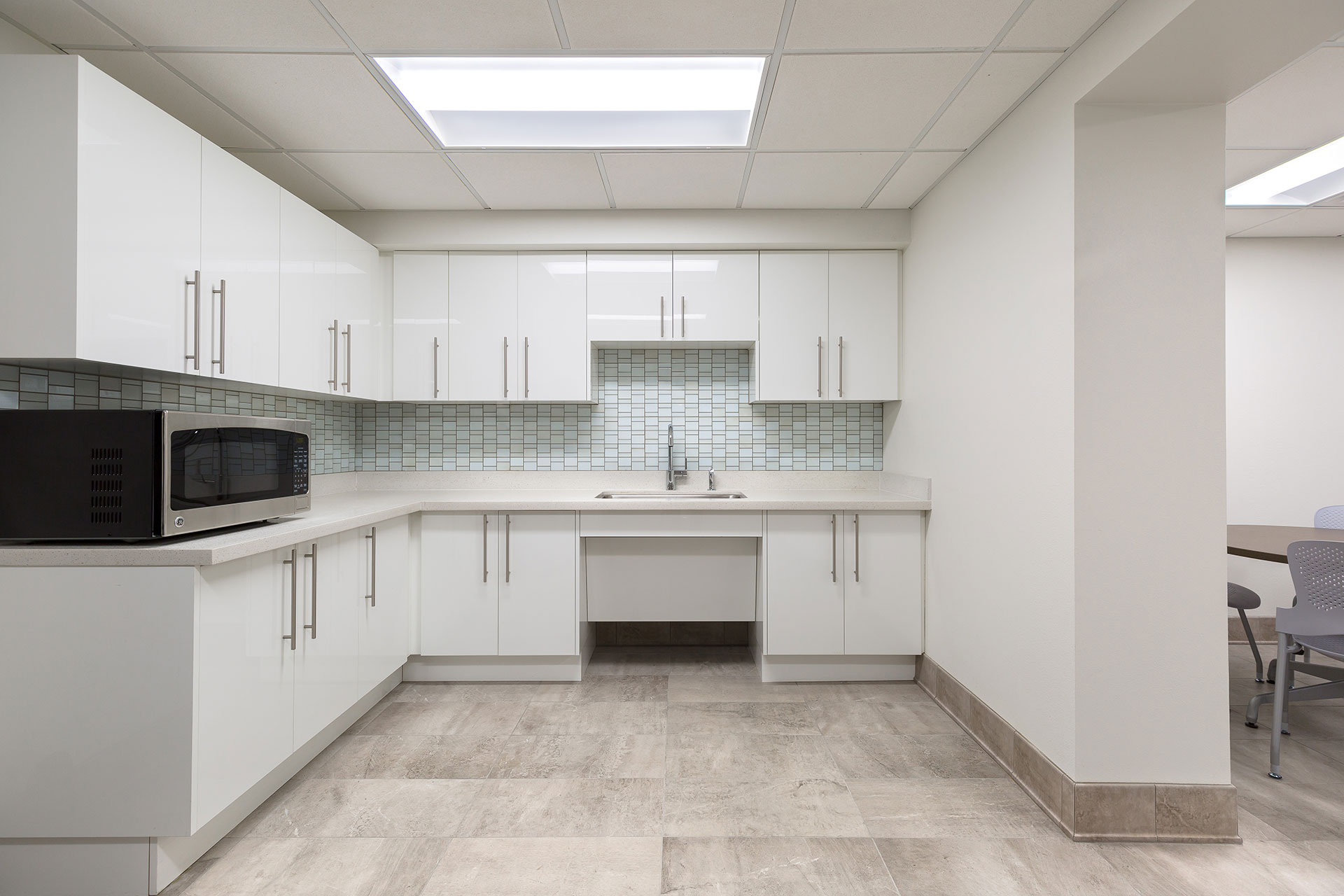 A white breakroom kitchenette with a white cabinets, a beige synthetic countertop, black microwave, and undermount sink, with a beige tile floor and paneled ceiling.