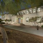 A white stucco two-story building at twilight with abundant windows exposing the interior and steps leading to the entrance like a dias with a mesquite tree in the foreground and desert landscaping about.