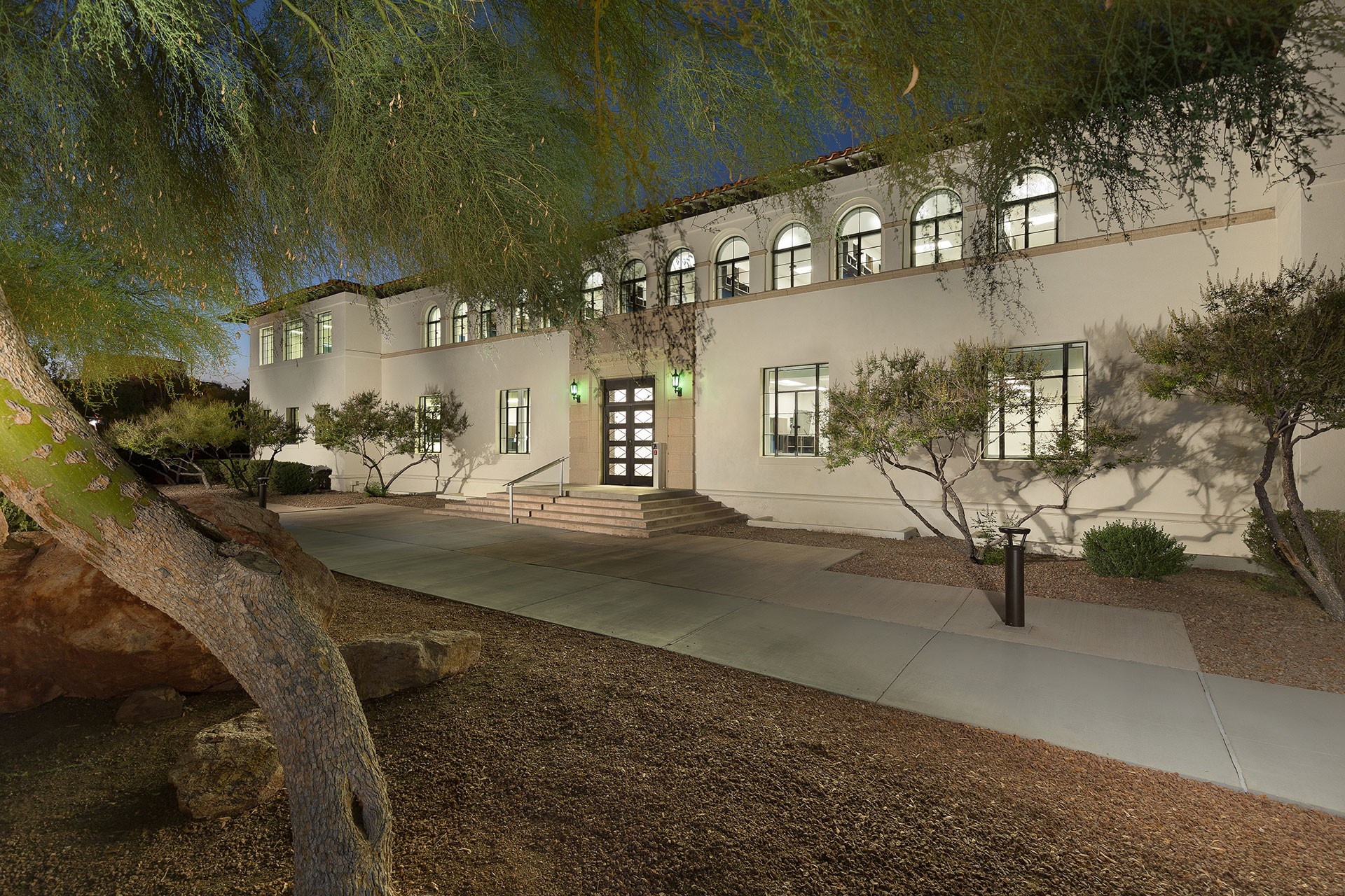 A white stucco two-story building at twilight with abundant windows exposing the interior and steps leading to the entrance like a dias with a mesquite tree in the foreground and desert landscaping about.