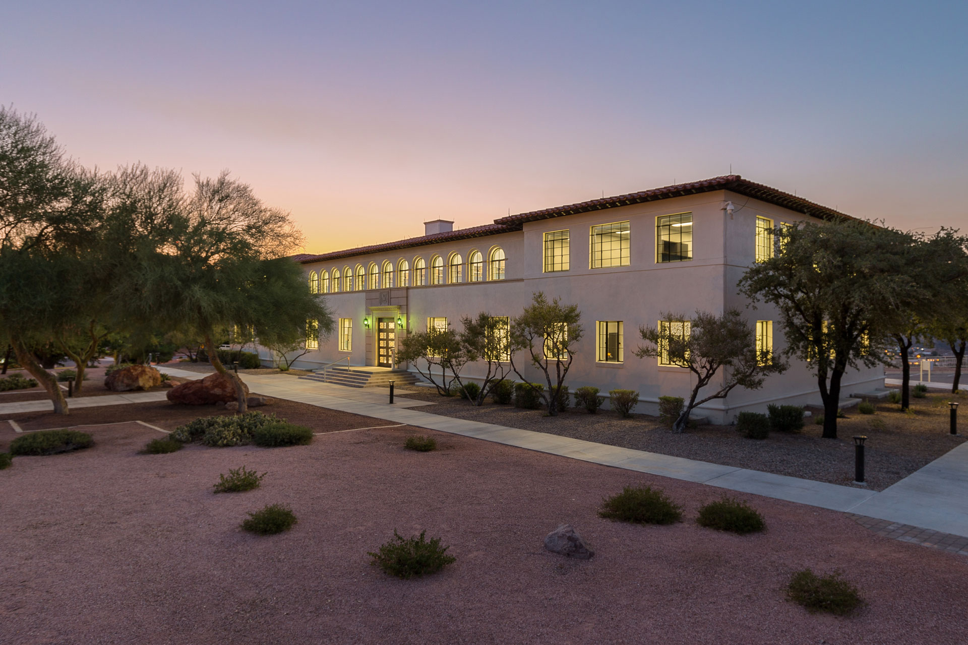 A white two-story stucco building with many windows and desert landscaping in the foreground and a yellow and purple sunset in the background.