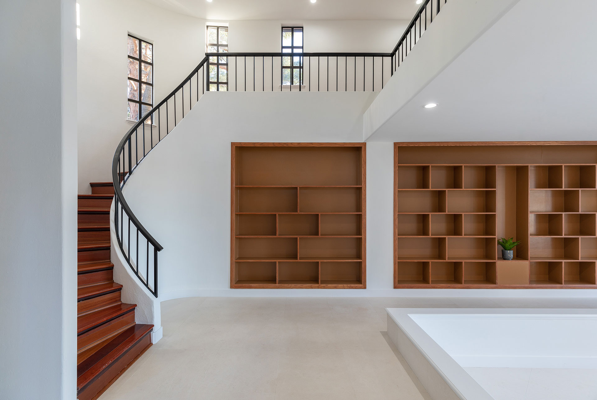 A white room with a brown built-in bookshelf with a spiral staircase wrapping around in with black railings.