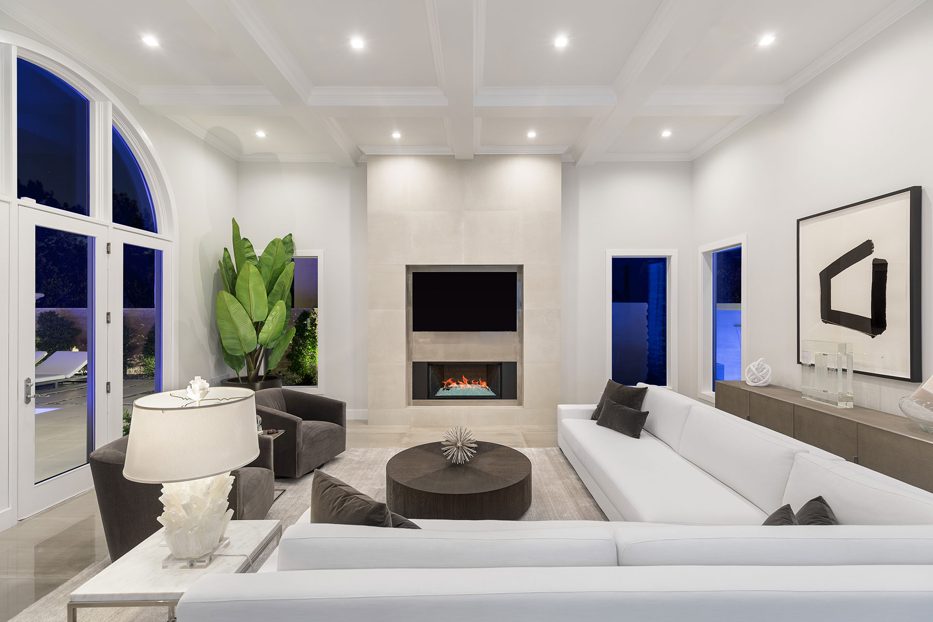 Living room with white walls and ceiling with a couch back in the foreground and a plat-panel tv and fireplace in the background, glass doors to patio chairs on the left and modern black-and-white art on the left wall.