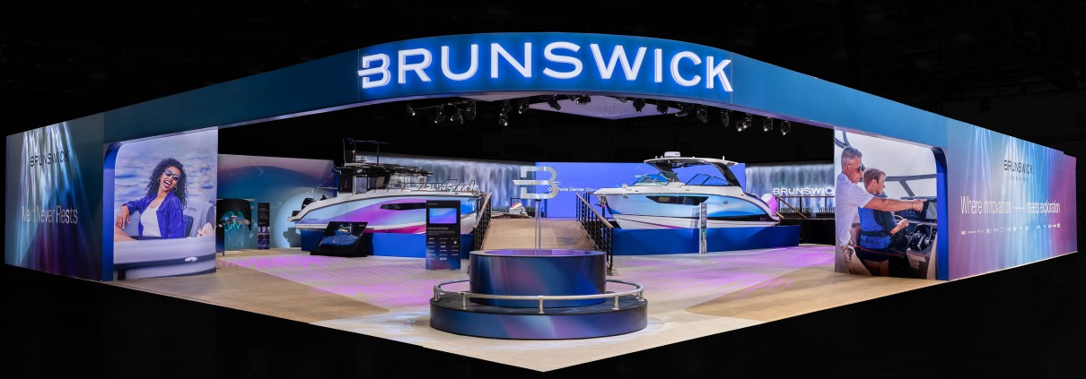 An exterior shot of the Brunswick booth in West Hall of the Las Vegas Convention Center at CES 2026. The exterior wall and archway entrance is blue with transitional gradients to white and purple with an illuminated Brunswick logo overhead. A circular pedestal with a silver B icon extending from the middle of a fountain is in the foreground. Two large blue, purple, and white boats are in the middle-ground on each side of a wooden-plank walkway up to an elevated platform. In the back are video walls with the Brunswick logo in the midst of a waterfall graphic. Everything but the Brunswick exhibit is darkened.