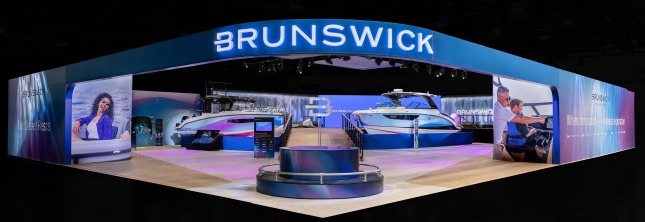 An exterior shot of the Brunswick booth in West Hall of the Las Vegas Convention Center at CES 2026. The exterior wall and archway entrance is blue with transitional gradients to white and purple with an illuminated Brunswick logo overhead. A circular pedestal with a silver B icon extending from the middle of a fountain is in the foreground. Two large blue, purple, and white boats are in the middle-ground on each side of a wooden-plank walkway up to an elevated platform. In the back are video walls with the Brunswick logo in the midst of a waterfall graphic. Everything but the Brunswick exhibit is darkened.