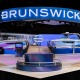 An exterior shot of the Brunswick booth in West Hall of the Las Vegas Convention Center at CES 2026. The exterior wall and archway entrance is blue with transitional gradients to white and purple with an illuminated Brunswick logo overhead. A circular pedestal with a silver B icon extending from the middle of a fountain is in the foreground. Two large blue, purple, and white boats are in the middle-ground on each side of a wooden-plank walkway up to an elevated platform. In the back are video walls with the Brunswick logo in the midst of a waterfall graphic. Everything but the Brunswick exhibit is darkened.