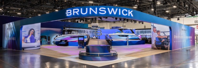 An exterior shot of the Brunswick booth in West Hall of the Las Vegas Convention Center at CES 2026. The exterior wall and archway entrance is blue with transitional gradients to white and purple with an illuminated Brunswick logo overhead. A circular pedestal with a silver B icon extending from the middle of a fountain is in the foreground. Two large blue, purple, and white boats are in the middle-ground on each side of a wooden-plank walkway up to an elevated platform. In the back are video walls with the Brunswick logo in the midst of a waterfall graphic.