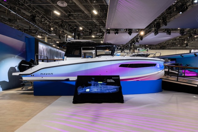 A profile of a white Navan C30 boat with a side panel in light blue with purple and magenta gradients at the Brunswick exhibit at CES 2026. The cabin roof is glossy black and a Fliteboard RACE is mounted on top. On the left, two black Mercury motors are visible at the rear of the boat. A ramp with leads to a raised platform behind the boat is visible on the right. A video wall is also visible in the background. Grey rectangular fabric tiles float above the exhibit amidst visible lighting rigging.