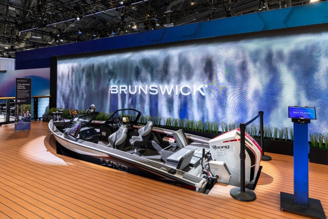 A Lund Crossover XS fishing boat with a Mercury Racing 150R outboard motor and Mercury Avator 35e kicker engine sits sunken into a wooden-plank platform at the Brunswick exhibit at CES 2026. Spot lighting accents the boat and a video wall displaying a waterfall pouring around a Brunswick logo is in the background. A small pedestal with an iPad stands on the right. Catwalks and lighting rigging is visible above.