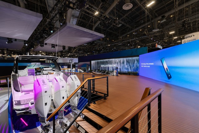 Stairs leading up to a raised wooden-plan platform in the Brunswick exhibit at CES 2026. On the right and leading to the back and center of the frame are two large video walls. On the left is a Sea Ray SLX 360 from back to front with its three white 425 V10 motors in the foreground. Two additional boats are visible in the background. Grey floating squares float above the exhibit with visible catwalks and lighting rigging around and behind them.