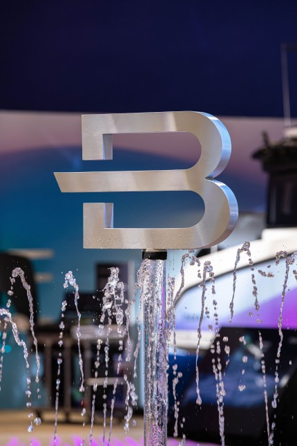 A close-up of an aluminum Brunswick "B" icon atop a glass rod at the Brunswick exhibit at CES 2026. Water shoots up from a surrounding fountain. The dark blue, white, light blue and purple hued exhibit walls as well as a boat with similar colors are out of focus in the background.
