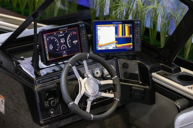 A close-up of the steering wheel and controls of a Lund fishing boat at the Brunswick exhibit at CES 2026. Two Lowrance monitors sit atop the steering wheel. The left-hand screen shows speed and fuel gauges. The right-hand screen show depth data and graphs. The console is black with silver and white gauges.